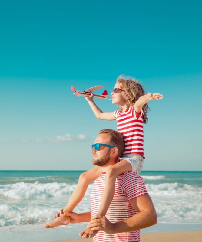 daughter holding toy airplane atop father's shoulders on beach